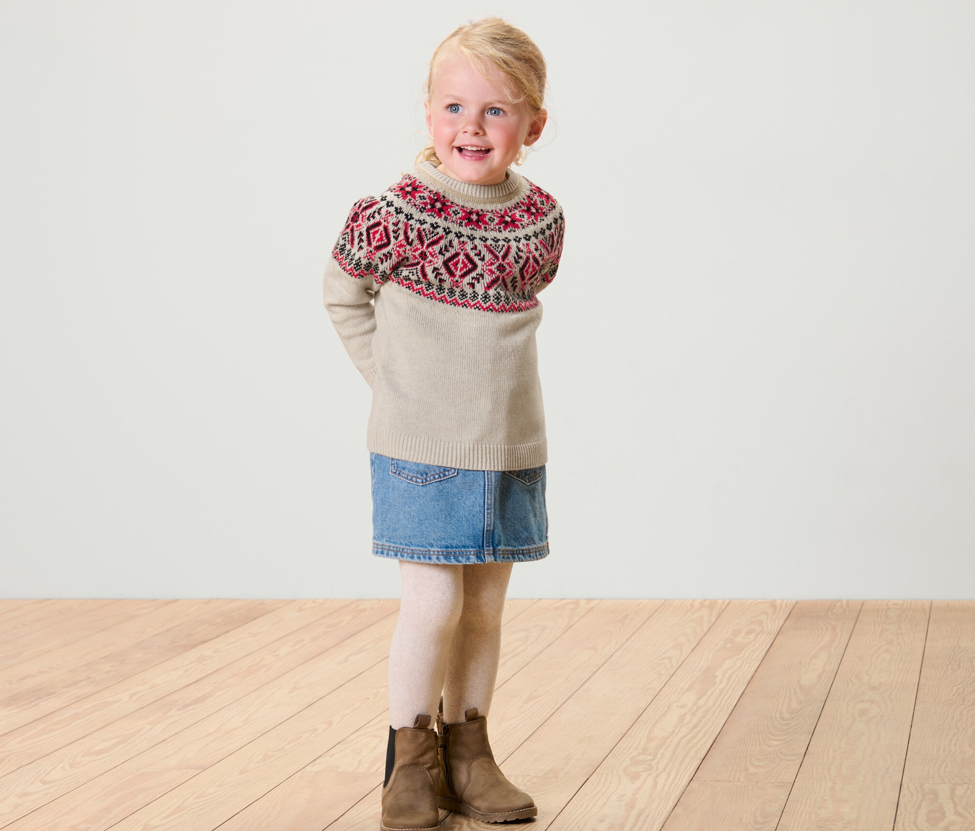 Fillette blonde souriante portant un pull norvégien enfant, une jupe en jean, des leggings blancs et des bottes marron.
