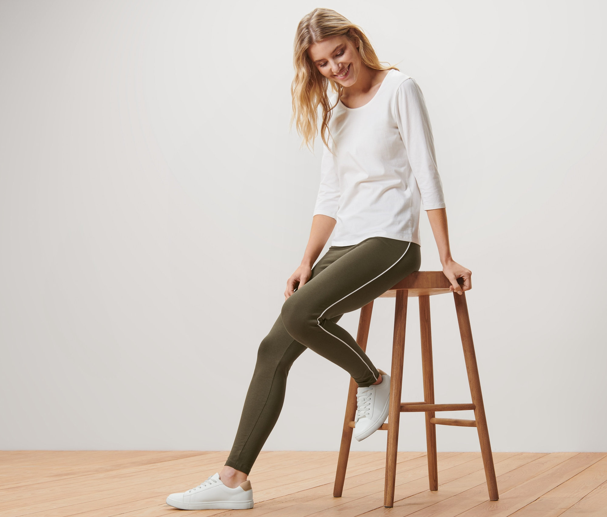 Une femme blonde est assise sur un tabouret en bois et porte un legging passepoilé vert, un t-shirt blanc et des baskets.