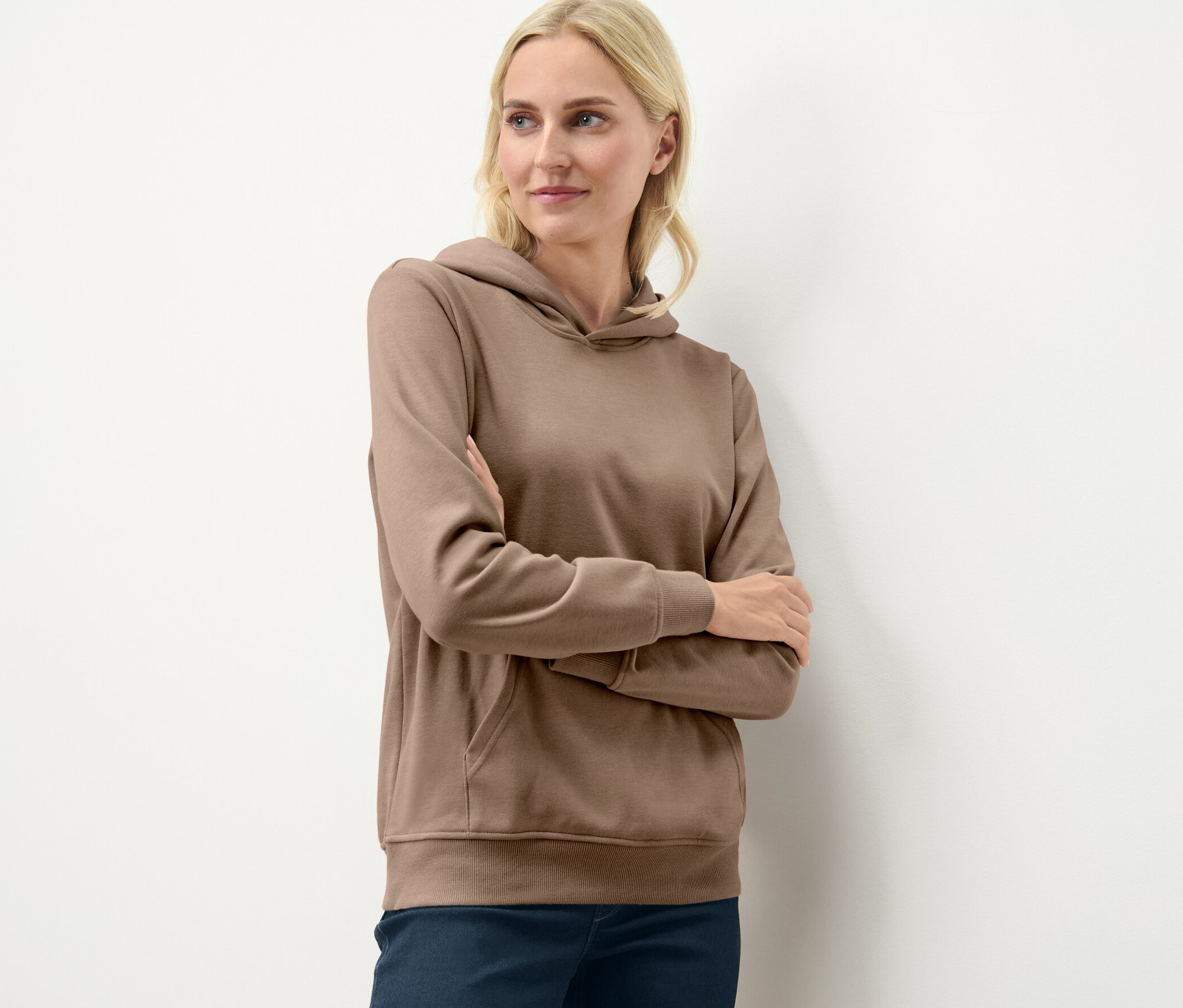 Une femme blonde pose les bras croisés, portant un sweat à capuche beige.