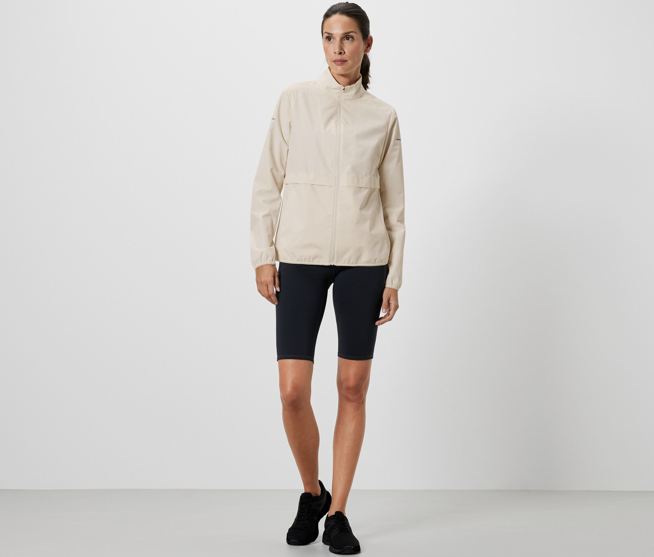 Une femme debout portant une veste de sport coupe-vent crème et un short de sport noir.