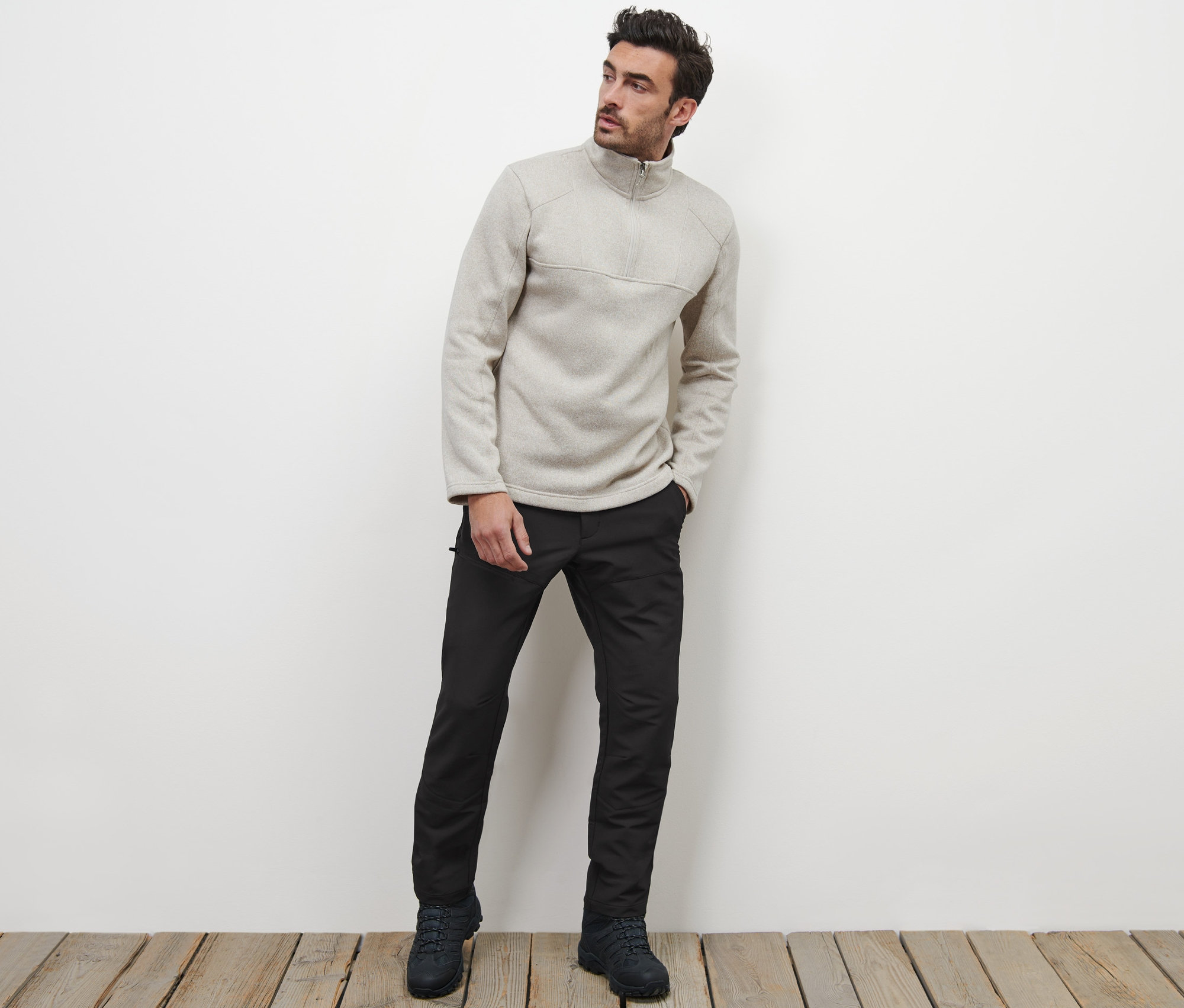 Un homme pose sur un plancher en bois, portant un pantalon thermique et un pull zippé.