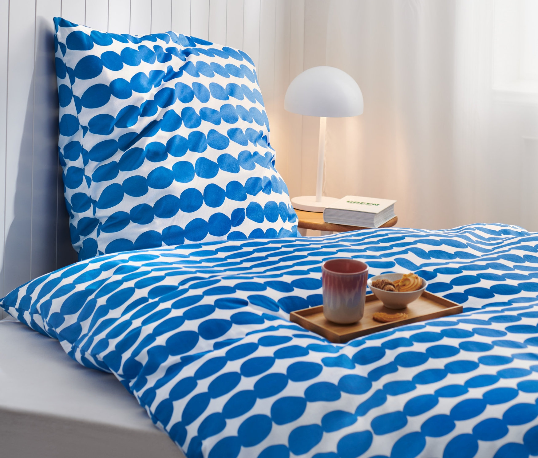 Gros plan d'un lit avec une parure de lit en coton de qualité supérieure foncée avec un motif ovale bleu. Un plateau avec une tasse et un bol de biscuits sont posés sur le lit.