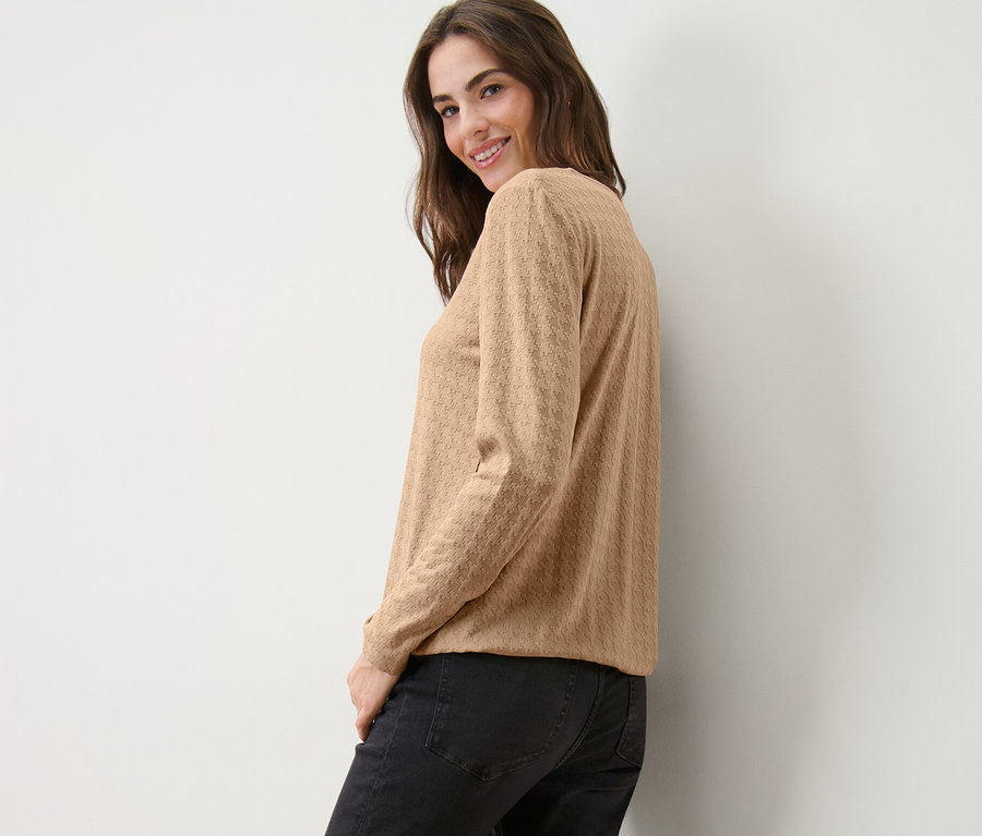 Une femme souriante regardant par-dessus son épaule pose, portant un t-shirt à manches longues structuré camel et un jean noir.