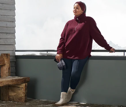Une femme appuiée contre une balustrade porte un pull oversize en peluche, un bandeau en maille rouge bordeaux, un legging thermique bleu et des bottines en cuir velours. Elle tient des moufles matelassées.