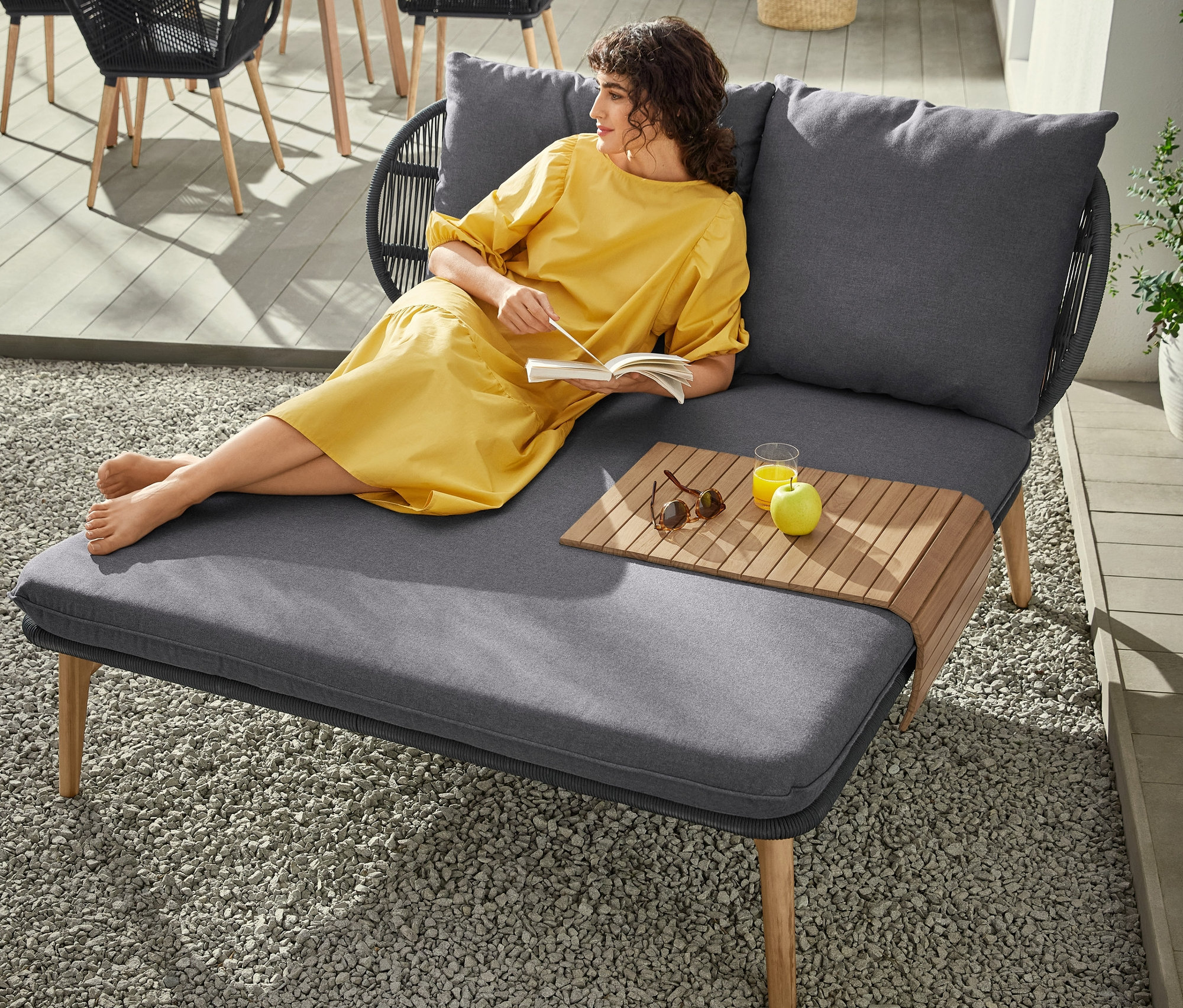 Une femme en robe jaune est allongée sur un lit de jour à partie tête en tressage textile et lit un livre. Sur la lit de jour il y a une table, une boisson et une pomme. À l'arrière-plan, il y a une table de jardin ronde, une chaise à coque avec cannage en textile et une lampe de table à LED à batterie.