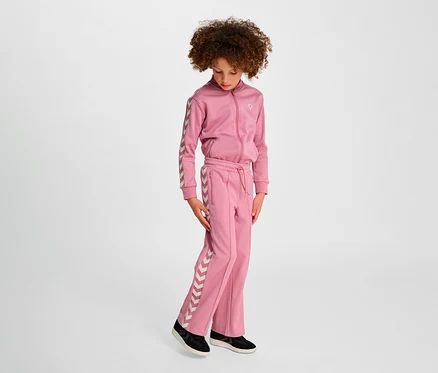 Une fille portant un survêtement rose Hummel HMLJR Archive Wideleg Poly Pants se tient sur un fond clair.