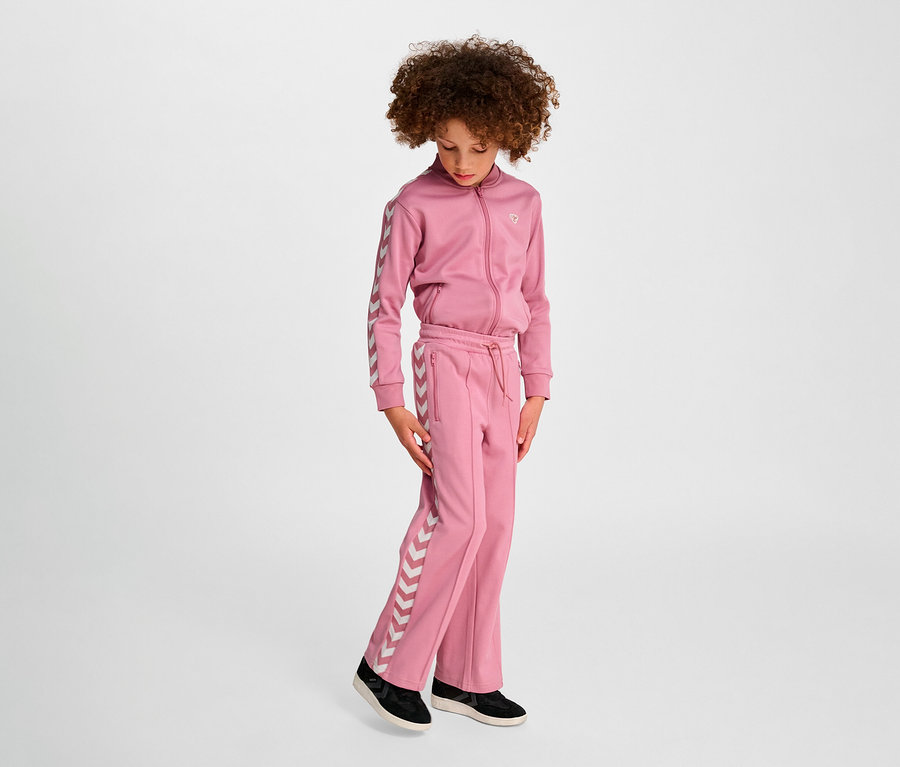 Une fille portant un survêtement rose Hummel HMLJR Archive Wideleg Poly Pants se tient sur un fond clair.