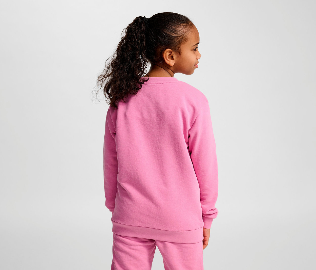 Fillette avec queue de cheval posant en survêtement rose. Elle porte un sweat-shirt HUMMEL HMLJR Base rose.