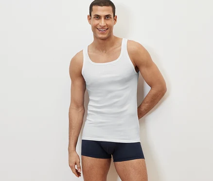 Un homme souriant pose sur un fond blanc, portant un des 2 hauts à doubles côtes blancs et un boxer bleu marine.