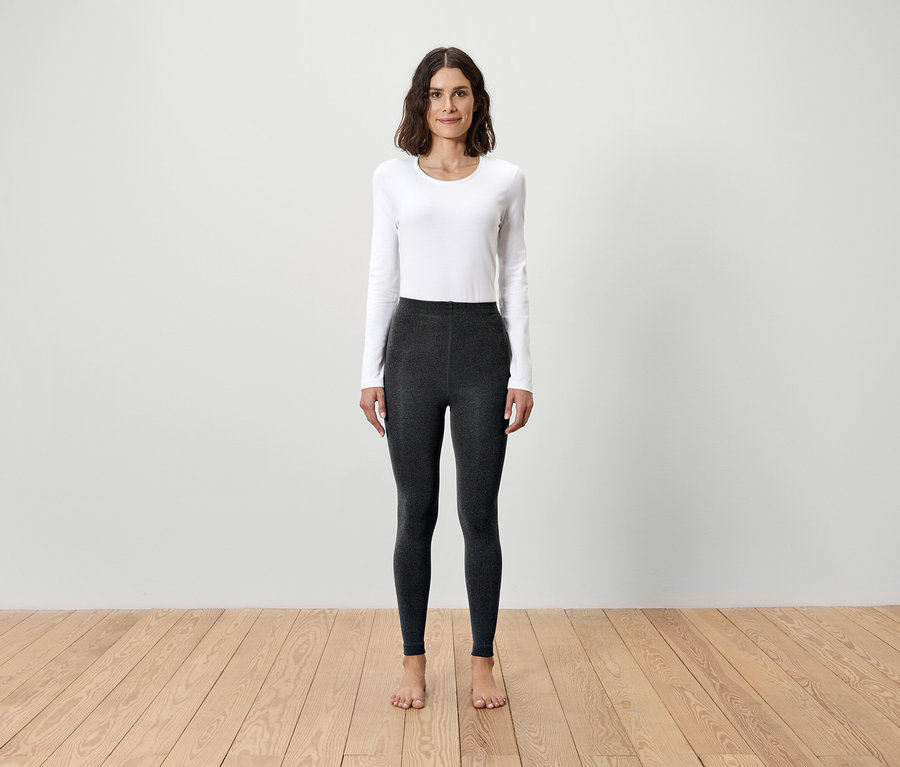 Une femme se tient pieds nus sur un plancher en bois, portant un t-shirt blanc à manches longues et un legging gris.