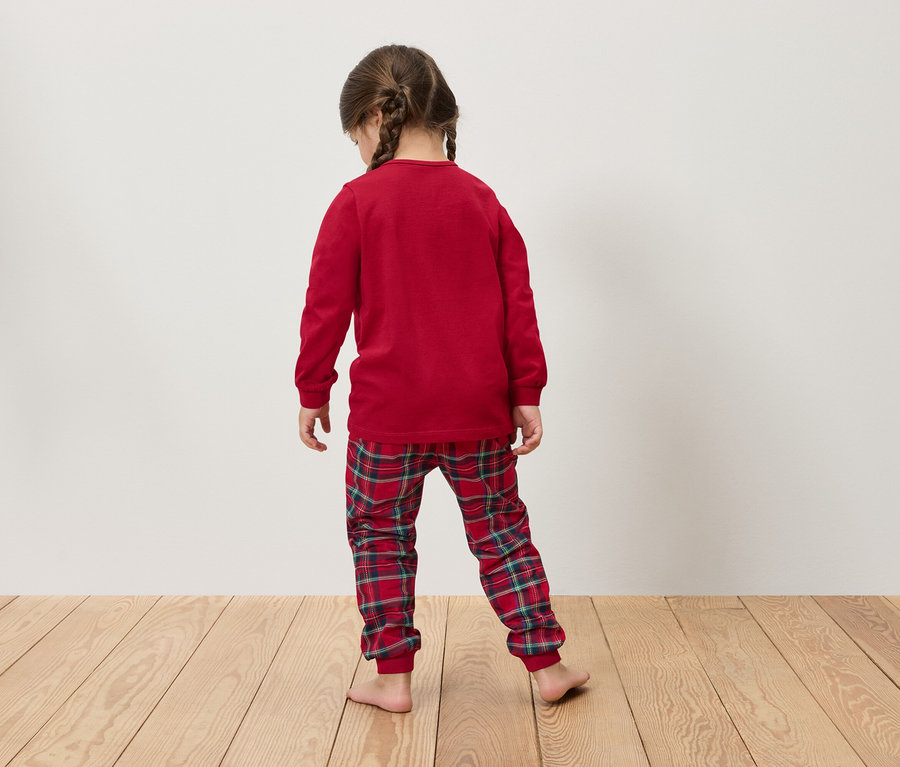 Une fillette portant un pyjama rouge à manches longues et un pantalon à carreaux se tient dos au spectateur sur un plancher en bois.