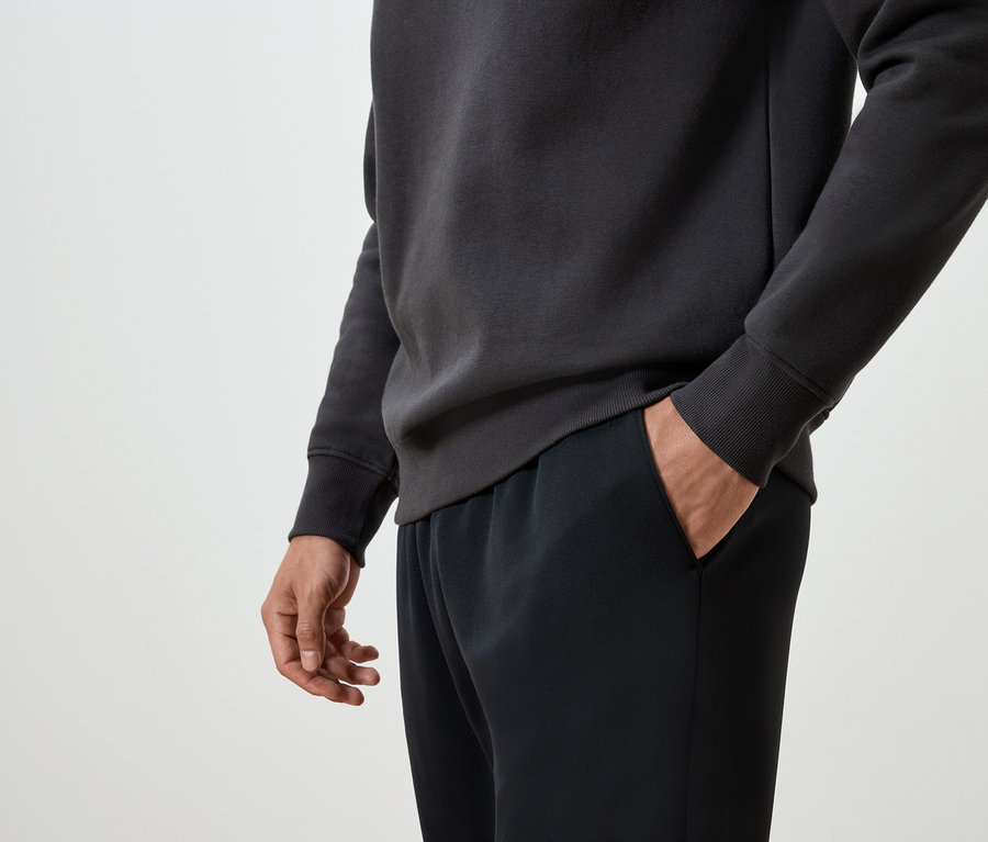 Un homme a une main dans la poche d'un pantalon de survêtement noir et porte un pull gris foncé.