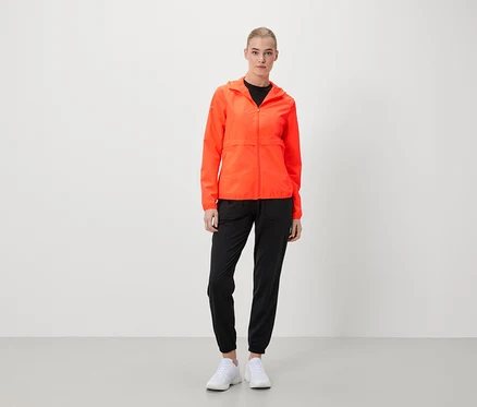 Une femme debout portant une veste orange, un pantalon noir et des chaussures blanches.