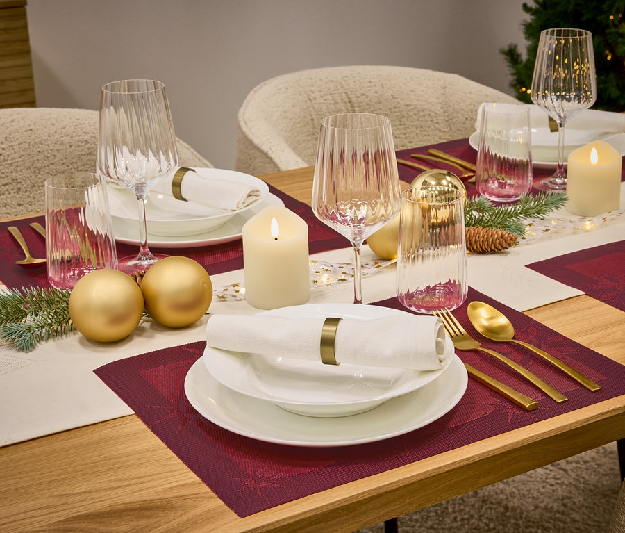 Une table dressée avec des sets de table bordeaux, de la vaisselle blanche, des verres, des couverts dorés, des bougies et des décorations.