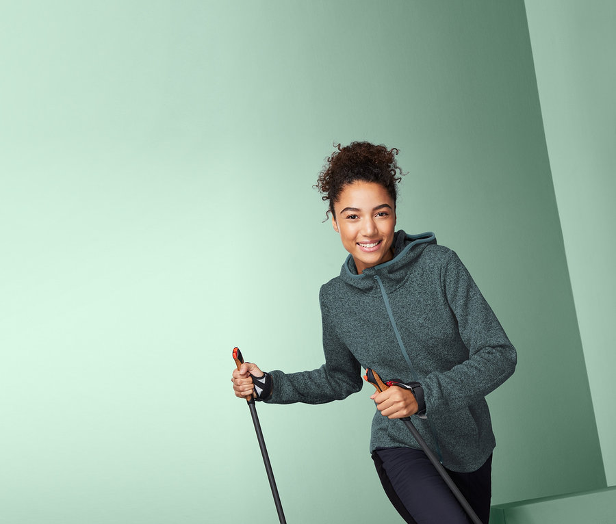 Femme souriante tenant des bâtons de randonnée et portant une veste à capuche en polaire tricotée.