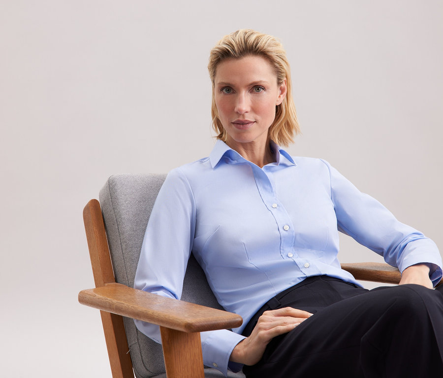 Une femme blonde est assise dans un fauteuil gris, portant une chemise bleue et un pantalon noir.