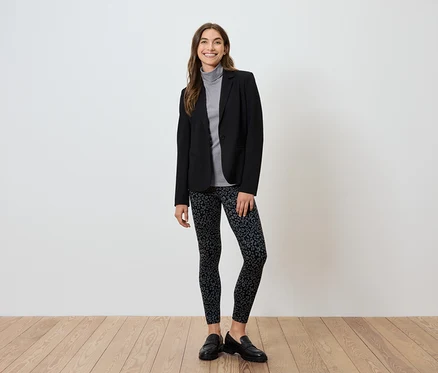 Femme en blazer noir, col roulé gris et leggings à motifs debout sur un plancher en bois.