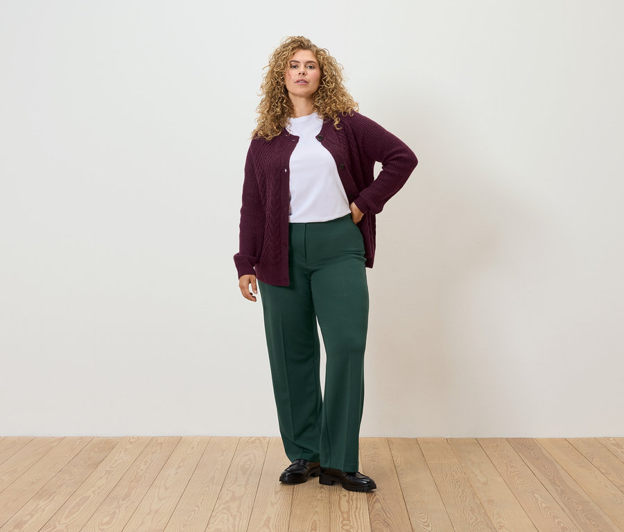 Une femme se tient debout, une main sur la hanche, portant un cardigan bordeaux, un t-shirt blanc et un pantalon vert.