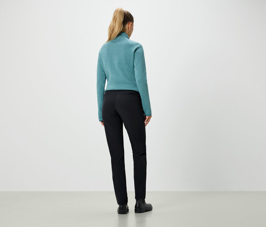 Vue arrière d'une femme blonde aux cheveux attachés en queue de cheval, portant un pull bleu-vert et un pantalon noir.