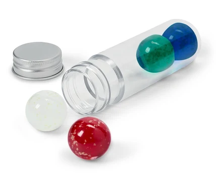 Un tube transparent est posé sur le côté avec une bille bleue et une bille verte à l'intérieur, un couvercle argenté à côté et une bille blanche et une bille rouge à l'extérieur.