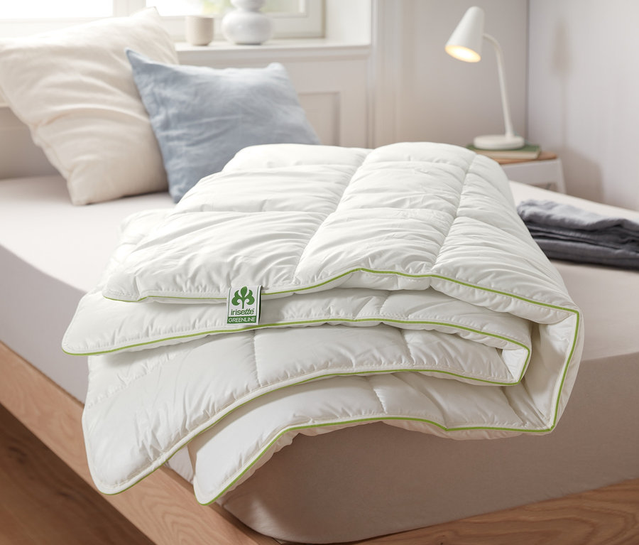 Une couette thermique irisette® greenline pliée avec une surface blanche et une bordure verte est posée sur un lit.