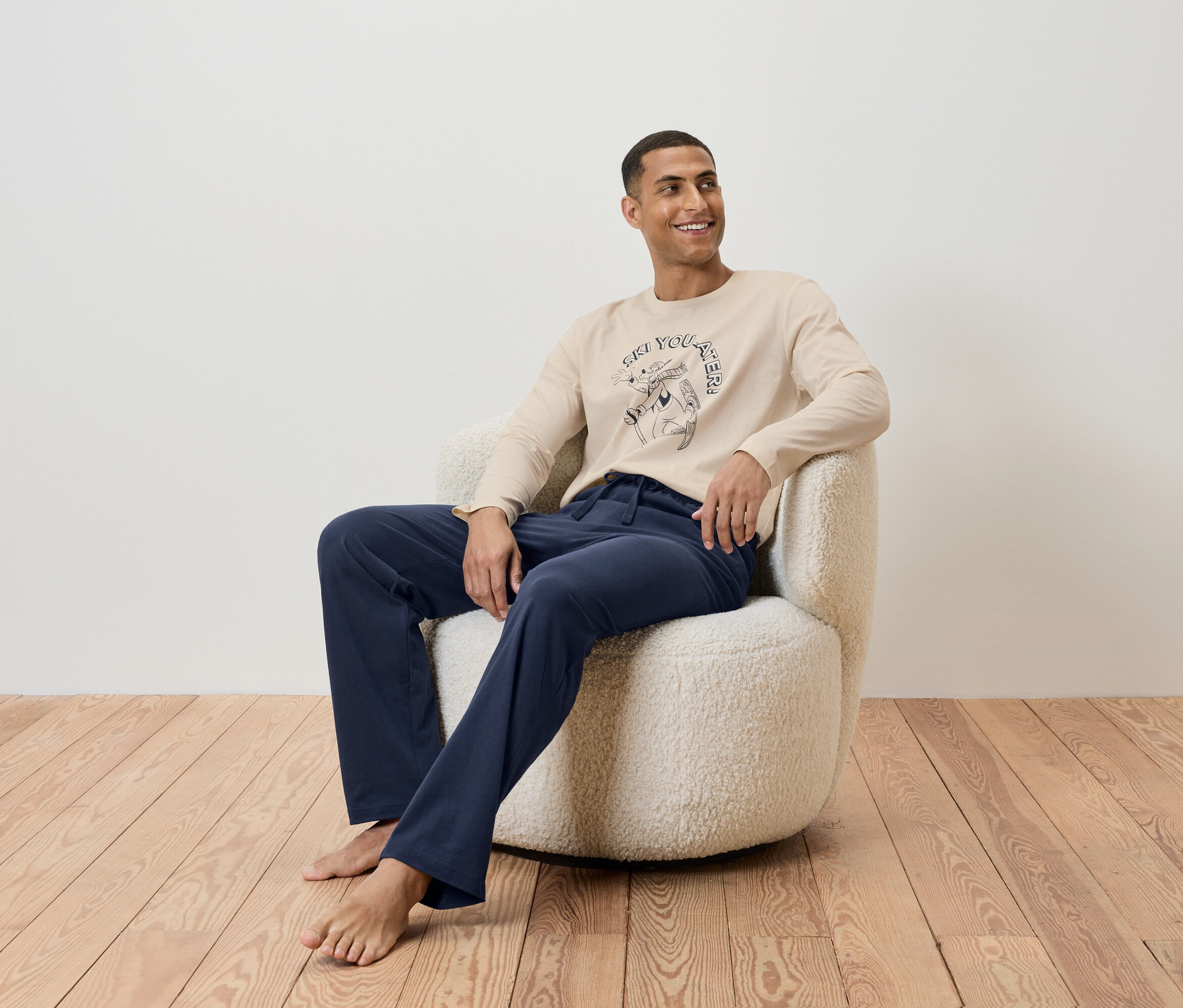 Un homme est assis dans un fauteuil et porte un t-shirt beige à manches longues et un pantalon bleu marine.