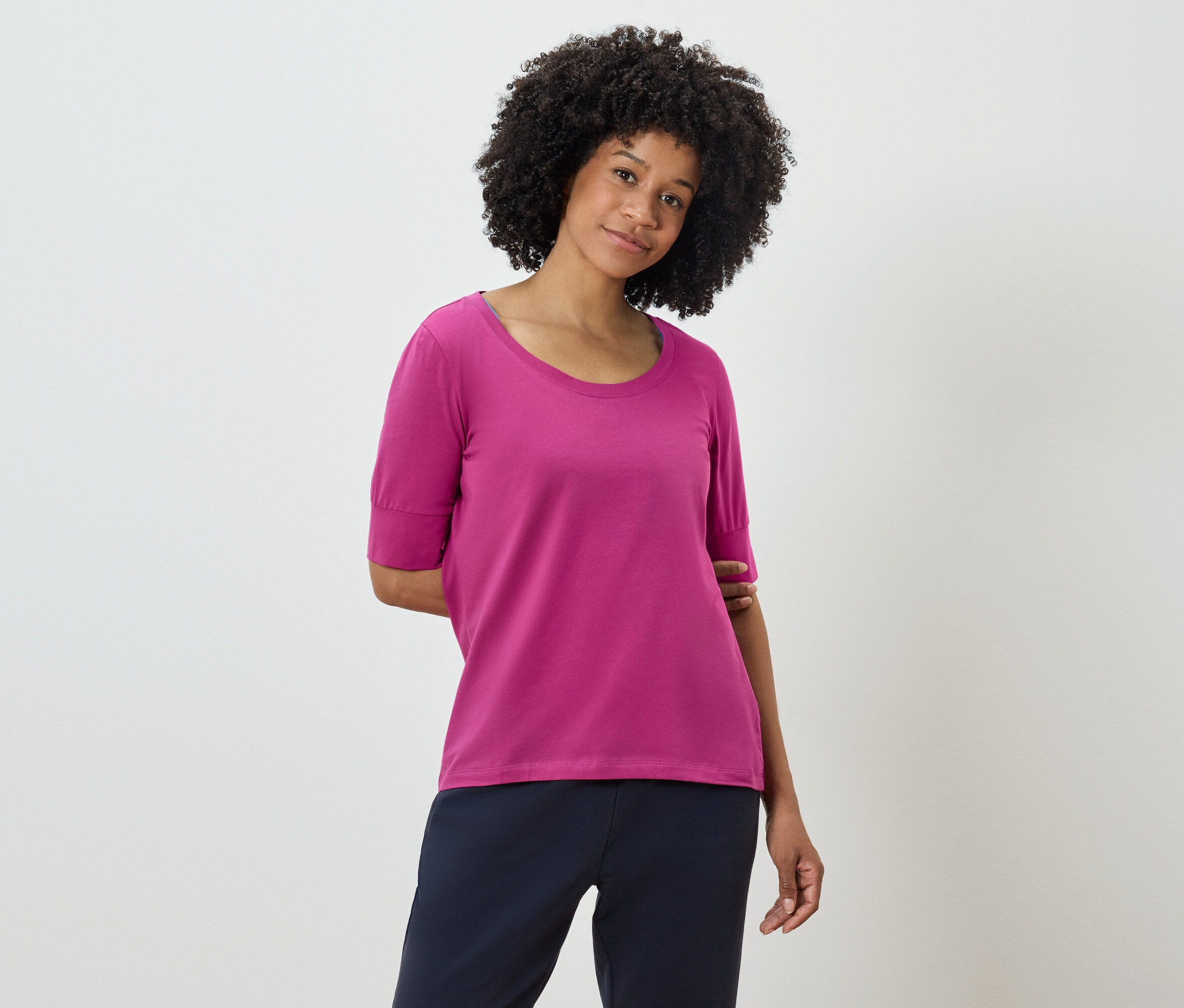 Femme avec coiffure afro posant dans un t-shirt rose et un pantalon bleu marine.
