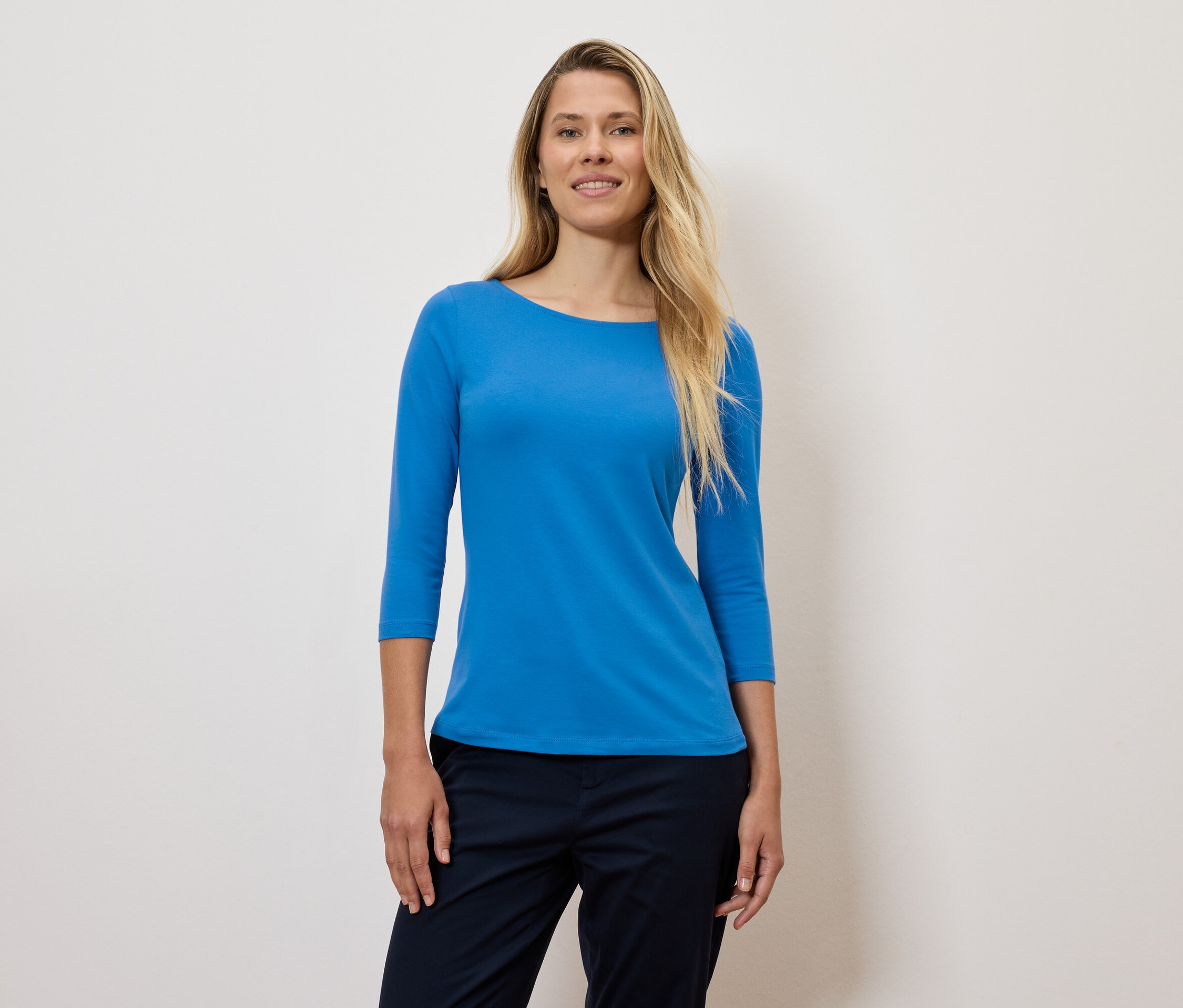 Une femme blonde porte un t-shirt bleu à manches trois-quarts et un pantalon bleu foncé.