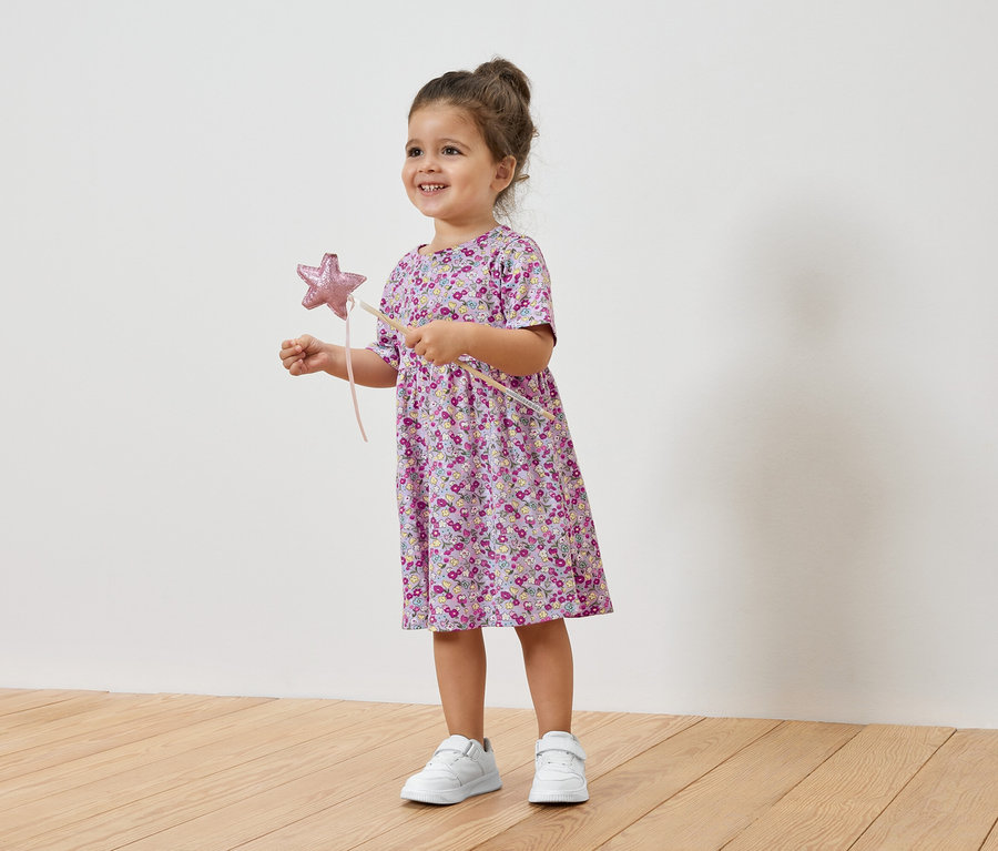 Petite fille souriante tenant une baguette étoilée, portant une robe à fleurs et des baskets.