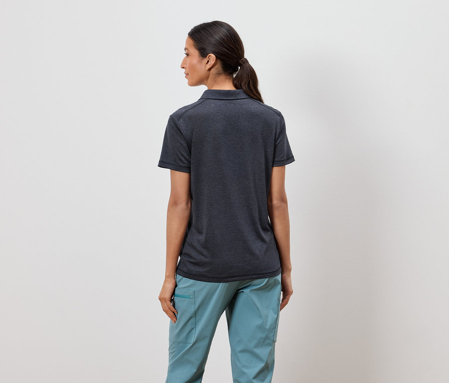 Une femme avec une queue de cheval se tient dos à la caméra, portant un polo gris foncé et un pantalon bleu clair.