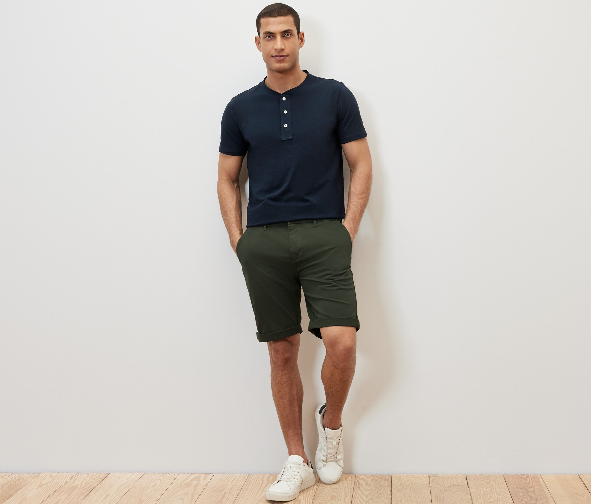 Un homme se tient debout, les mains dans les poches de son short. Il porte un Henley à manches courtes bleu marine.