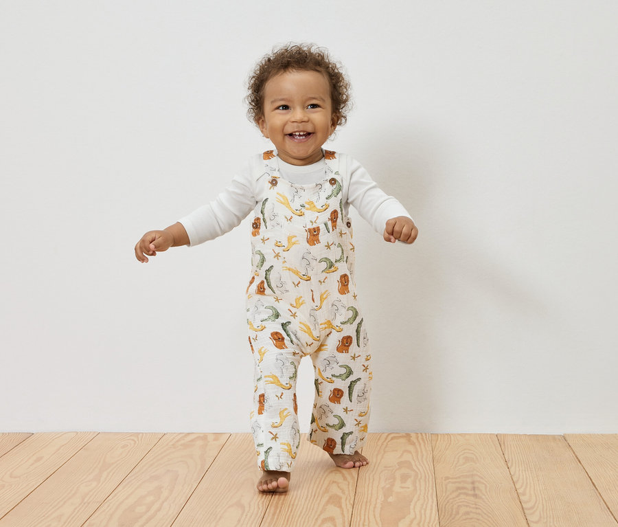 Un bébé souriant aux cheveux bouclés se tient sur un plancher en bois, portant un haut blanc à manches longues sous une salopette blanche à imprimé animal.