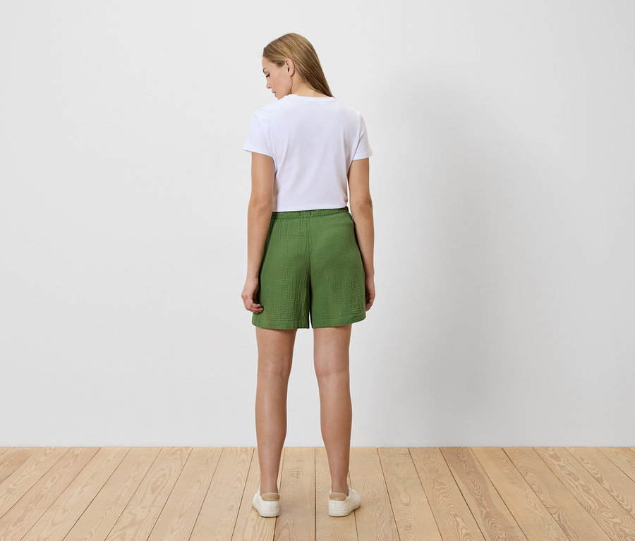 Vue arrière d'une femme portant un t-shirt blanc et un short vert, debout sur un plancher en bois.