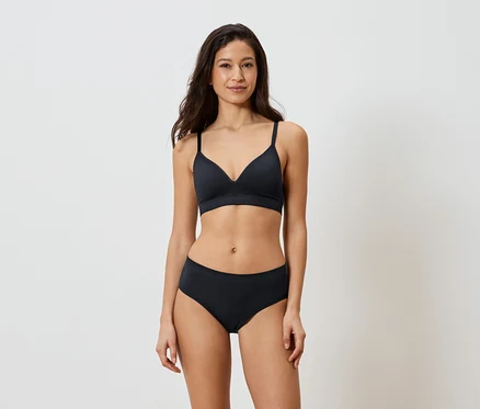 Femme aux cheveux foncés portant un soutien-gorge et une culotte noirs sur fond blanc.