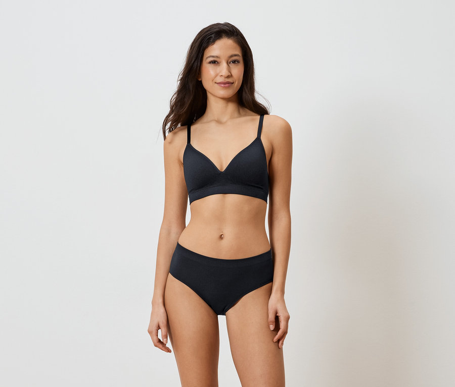 Femme aux cheveux foncés portant un soutien-gorge et une culotte noirs sur fond blanc.