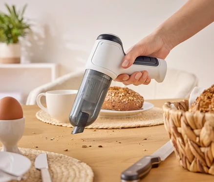 Une main aspire des miettes avec un aspirateur à main blanc sur une table en bois. Il y a aussi un œuf, une tasse et du pain sur la table.