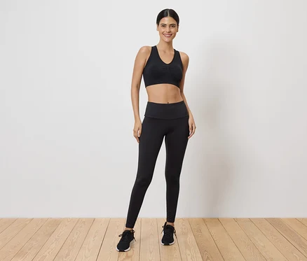 Une femme portant un soutien-gorge de sport noir et des leggings, debout sur un plancher en bois.