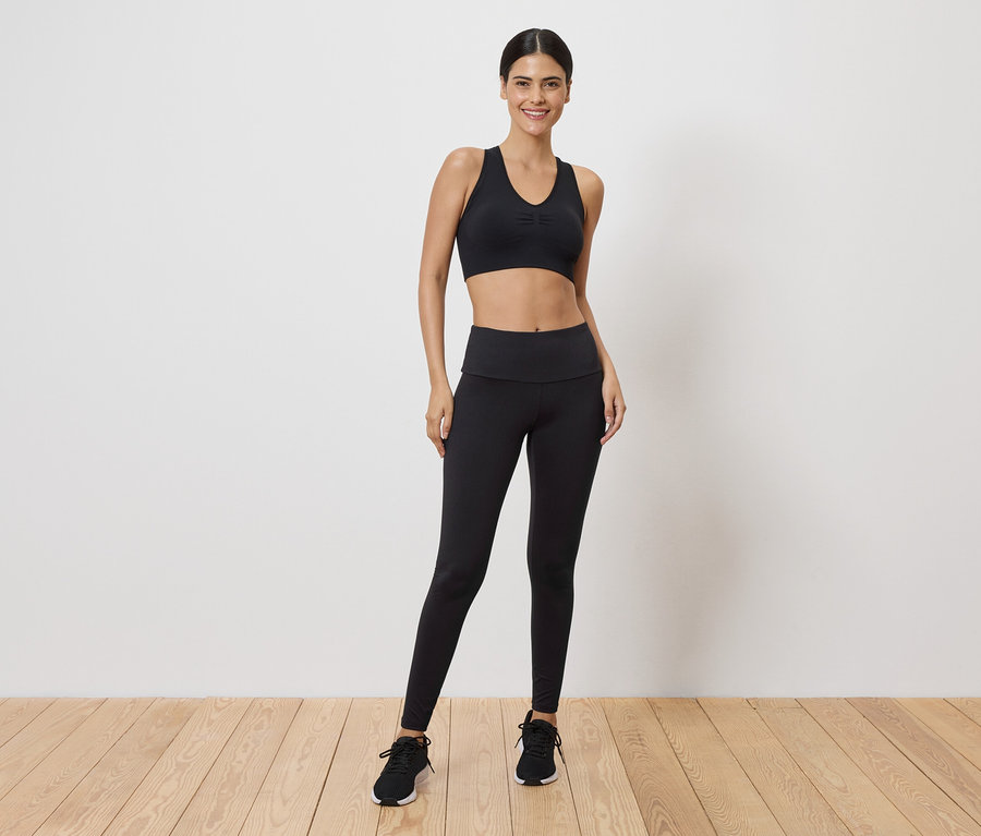 Une femme portant un soutien-gorge de sport noir et des leggings, debout sur un plancher en bois.