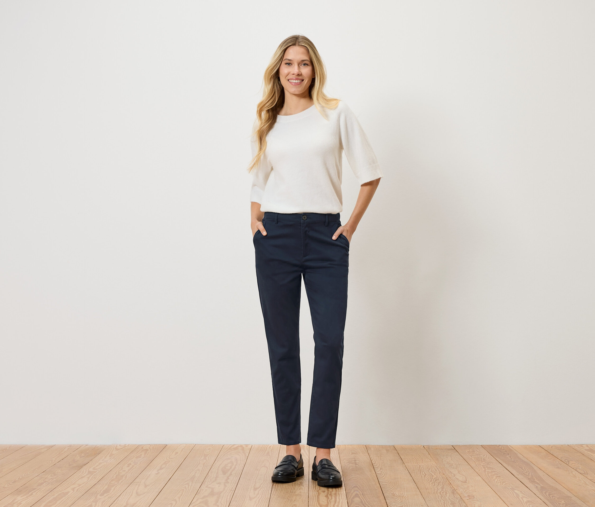 Une femme aux cheveux blonds ondulés se tient debout, les mains dans les poches de son pantalon. Elle porte un pull blanc, un pantalon bleu marine et des chaussures noires.