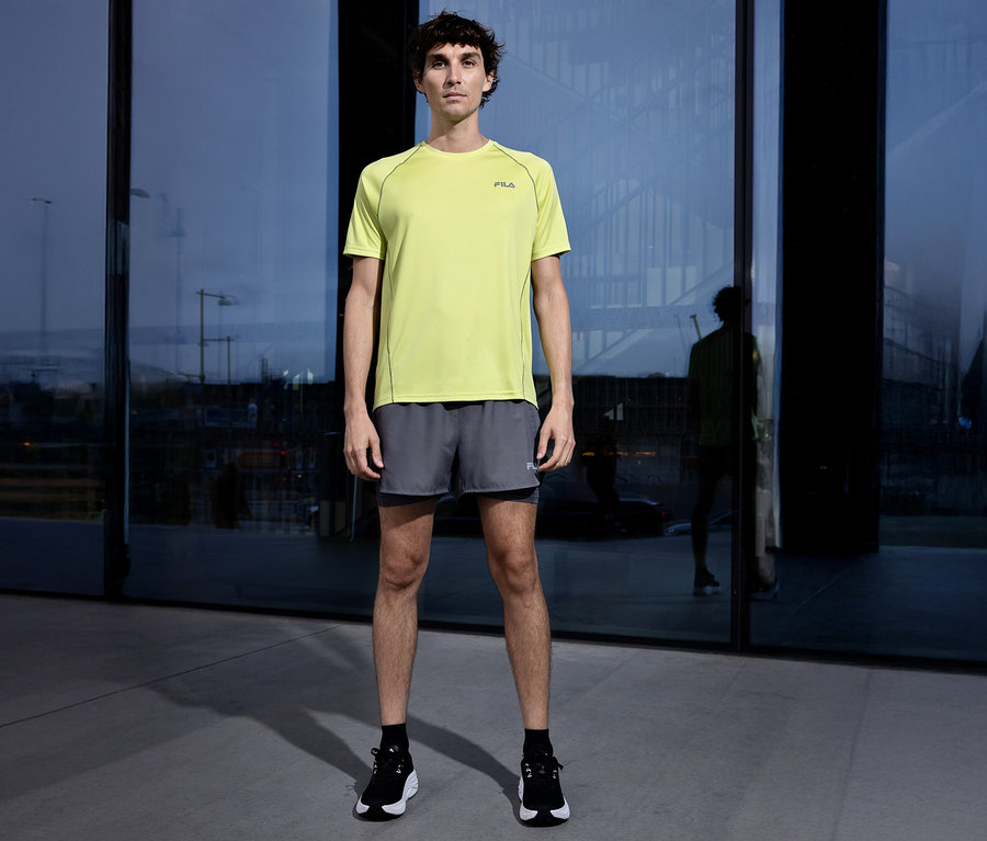 Un homme se tient devant un immeuble, portant un t-shirt Fila jaune, un short gris et des chaussures noires.