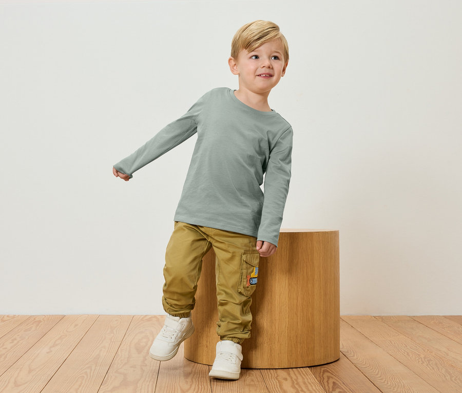 Un garçon blond portant un t-shirt vert à manches longues et un pantalon kaki se tient à côté d'un tabouret en bois.