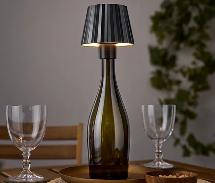 Une lampe de table en forme de bouteille de vin est posée sur une table en bois à côté de deux verres à vin et d'un bol de noix.