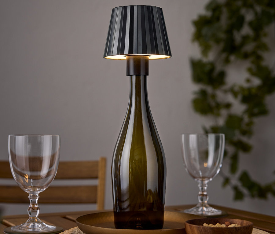 Une lampe de table en forme de bouteille de vin est posée sur une table en bois à côté de deux verres à vin et d'un bol de noix.