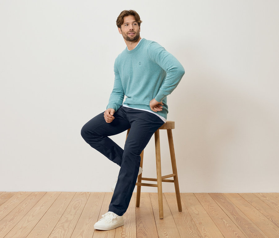 Un homme est assis sur un tabouret, portant un pull menthe, un pantalon bleu marine et des baskets blanches.