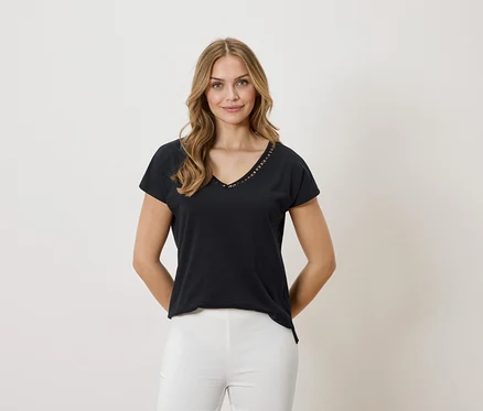 Une femme blonde porte un t-shirt noir à col en V et un pantalon blanc sur un fond neutre.