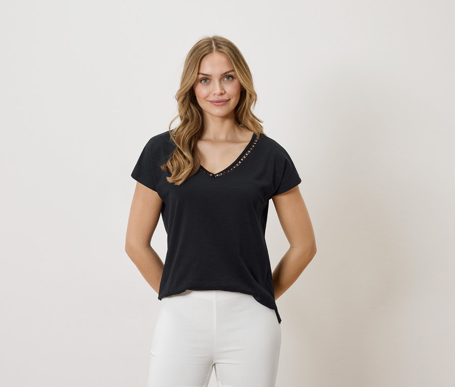 Une femme blonde porte un t-shirt noir à col en V et un pantalon blanc sur un fond neutre.