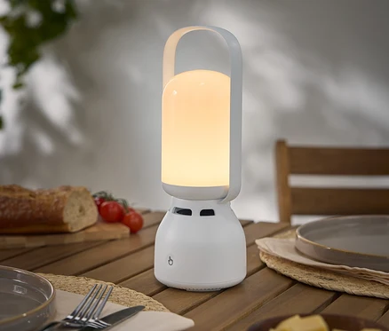 Une lampe blanche portable allumée avec une poignée est posée sur une table en bois à côté d'une assiette, de couverts, de pain, de tomates et de fromage.