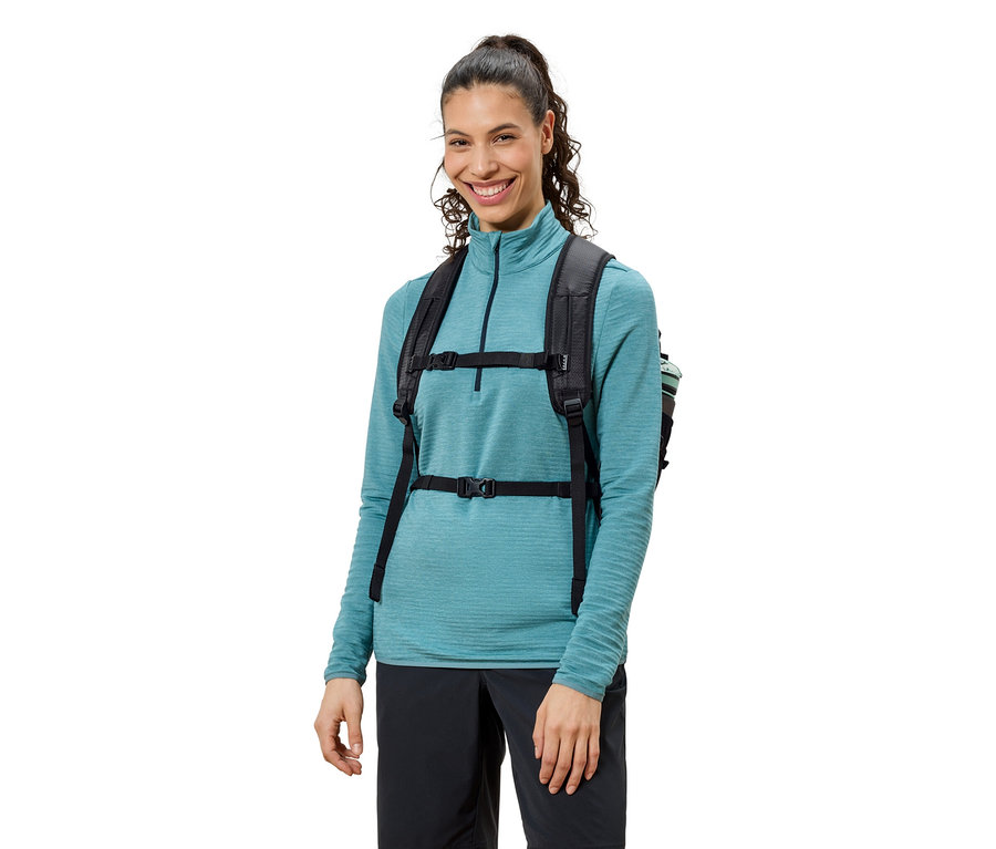 Femme souriante portant un sac à dos noir, un haut bleu et un pantalon noir.