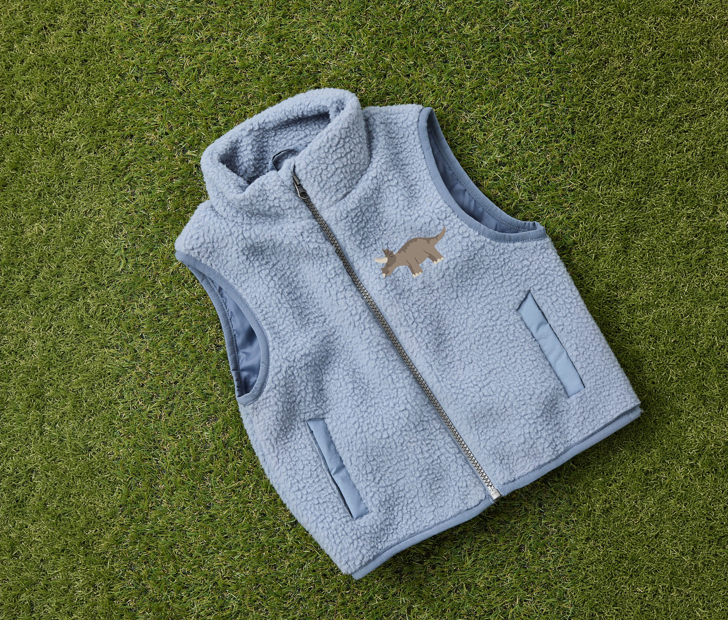 Un gilet en teddy pour enfant NAME IT bleu clair avec fermeture éclair et motif de dinosaure posé sur l'herbe.
