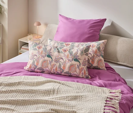 Une chambre avec un lit orné d'une housse de couette violette, d'oreillers à motifs floraux et d'une couverture crème à franges.